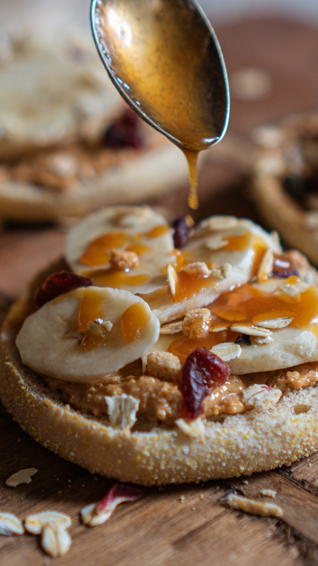 Close up of breakfast muffin with nuts, almonds, banana slices and herbal honey drizzled on top on a wooden surface.