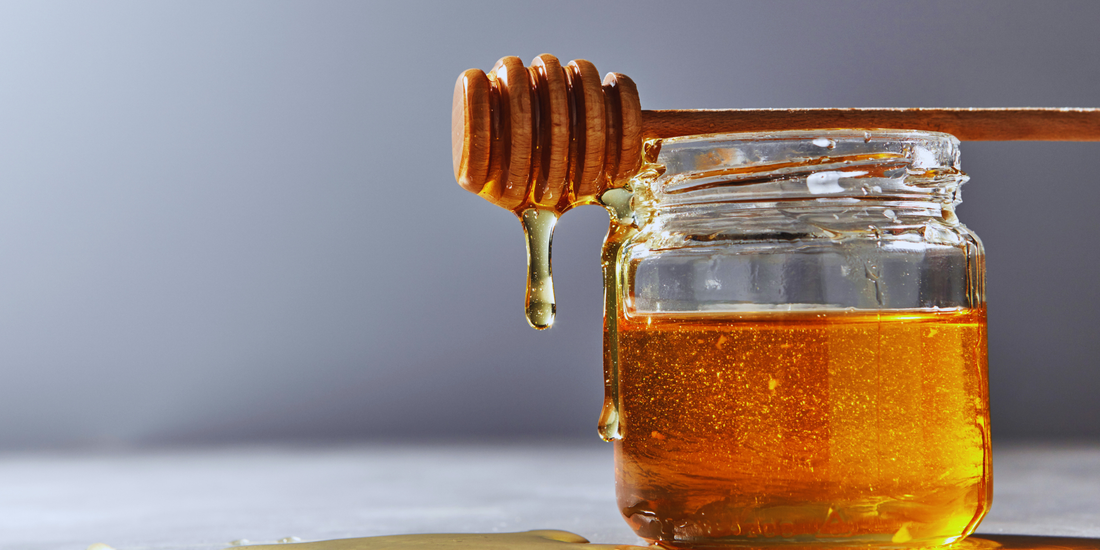 A clear jar of honey with a honey dipper sitting on top of it with honey dripping from it.