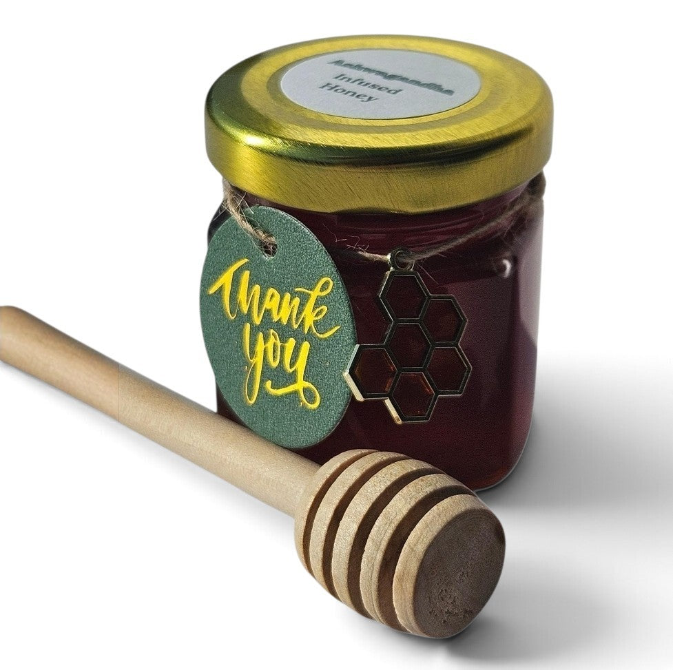 A jar of ashwagandha infused honey with a gold lid and a green 'thank you' tag, shown with a wooden honey dipper on a white background.