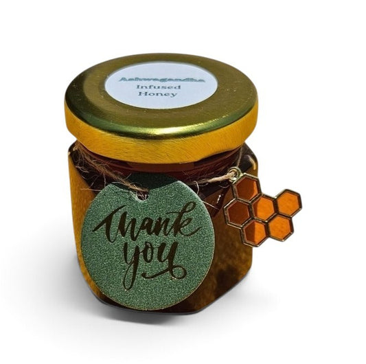 A small glass jar of infused honey with a gold lid, featuring a green glitter heart-shaped tag that says 'thank you' and a honeycomb charm, on a white background.