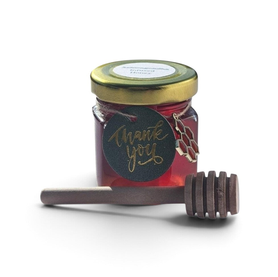 A small glass jar filled with red honey, topped with a gold lid and a round black 'thank you' tag, with a wooden honey dipper placed in front on a white background.
