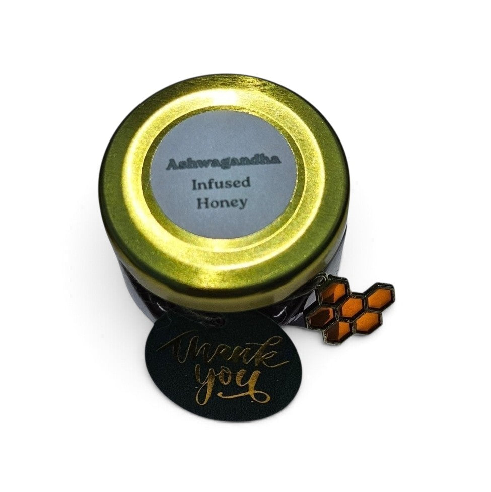 A small round tin of Ashwagandha Infused Honey with a metallic gold lid and a gray label, shown on a white background with a black 'Thank you' tag and an orange honeycomb charm.