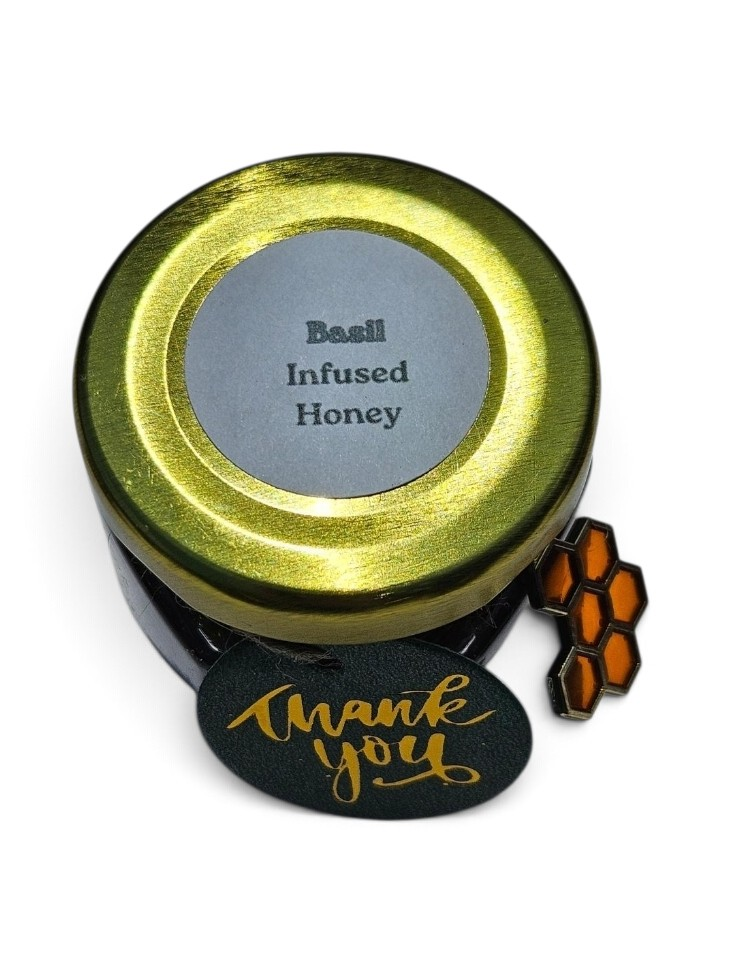 A round metal tin of Basil Infused Honey with a shiny gold lid and a gray label, shown on a white background with a black 'thank you' tag and an orange honeycomb enamel pin.