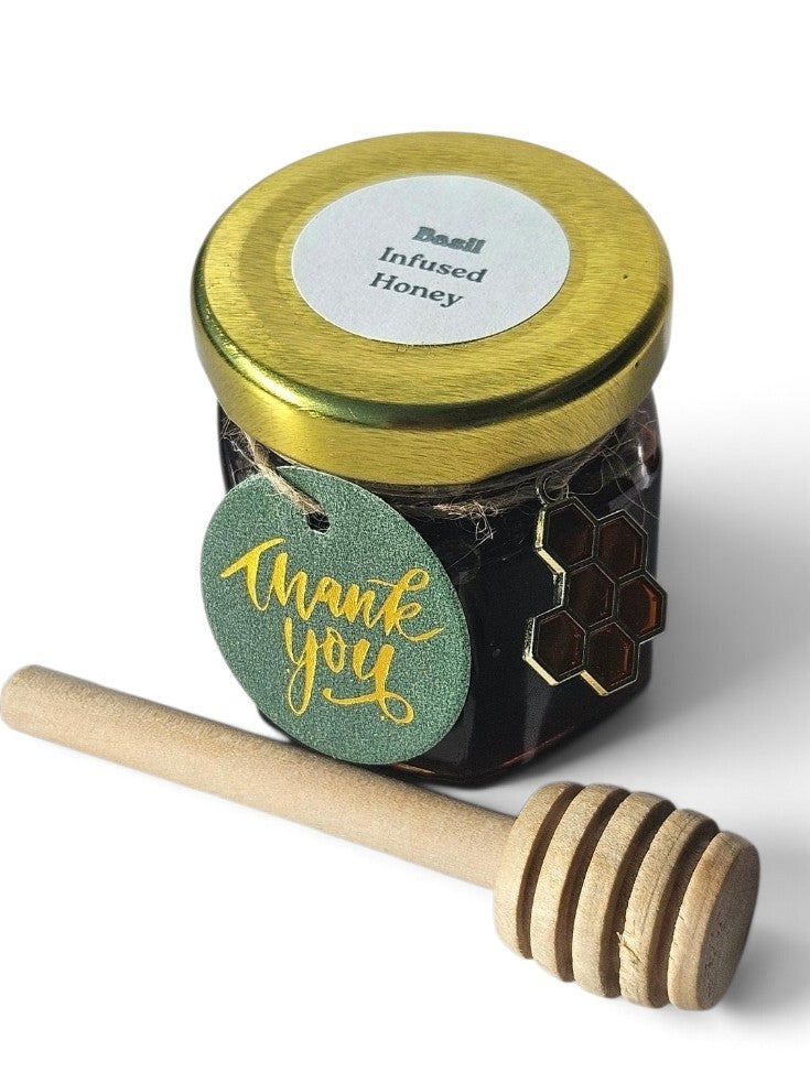 A small jar of basil infused honey with a gold lid and a green 'thank you' tag, shown with a wooden honey dipper on a white background.