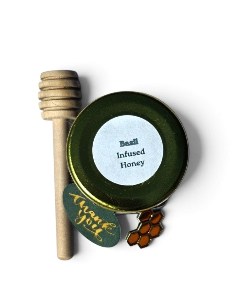A small round tin of infused honey with a green lid and white label reading 'Basil Infused Honey,' shown with a wooden honey dipper and a small honeycomb charm on a white background.