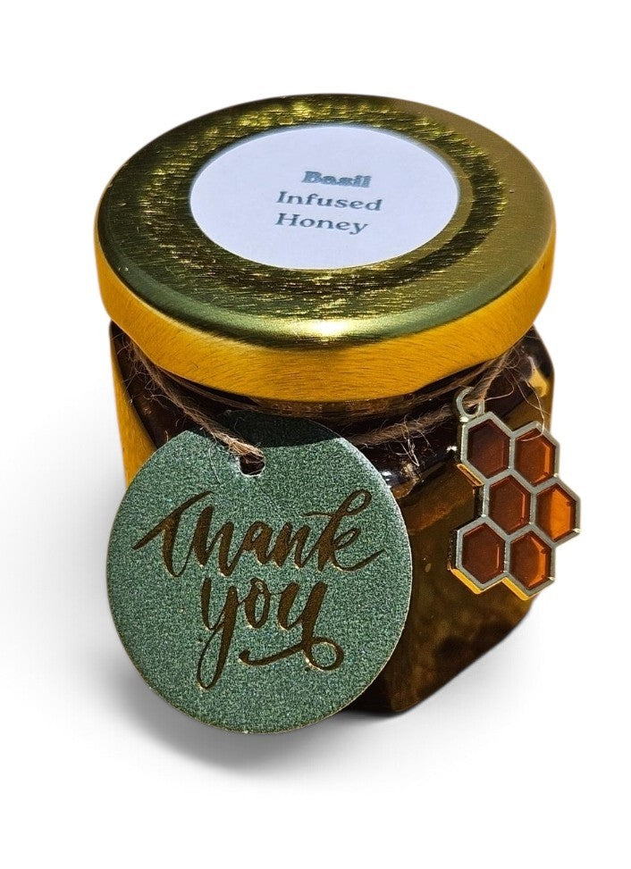 A jar of basil infused honey with a gold lid, a round green 'thank you' tag, and a honeycomb charm, shown on a white background.