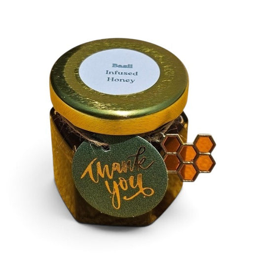 A small jar of infused honey with a gold foil lid and a round green 'thank you' tag attached, shown on a plain white background.