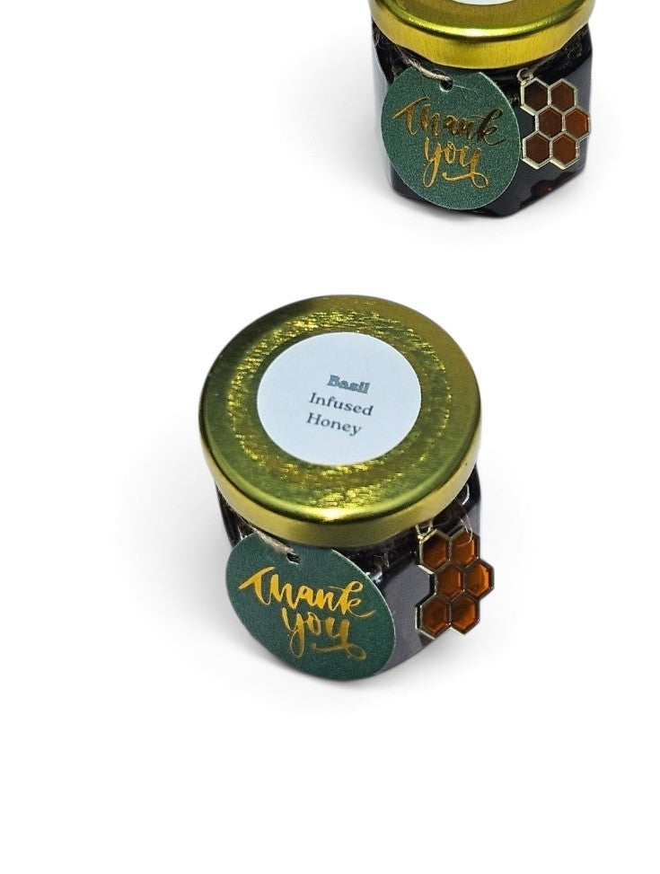 Two small jars of infused honey on a white background, one in the foreground and one partially visible in the background. The jars have gold lids, dark glass bodies, and round green labels with gold script reading 'thank you' and a honeycomb design.