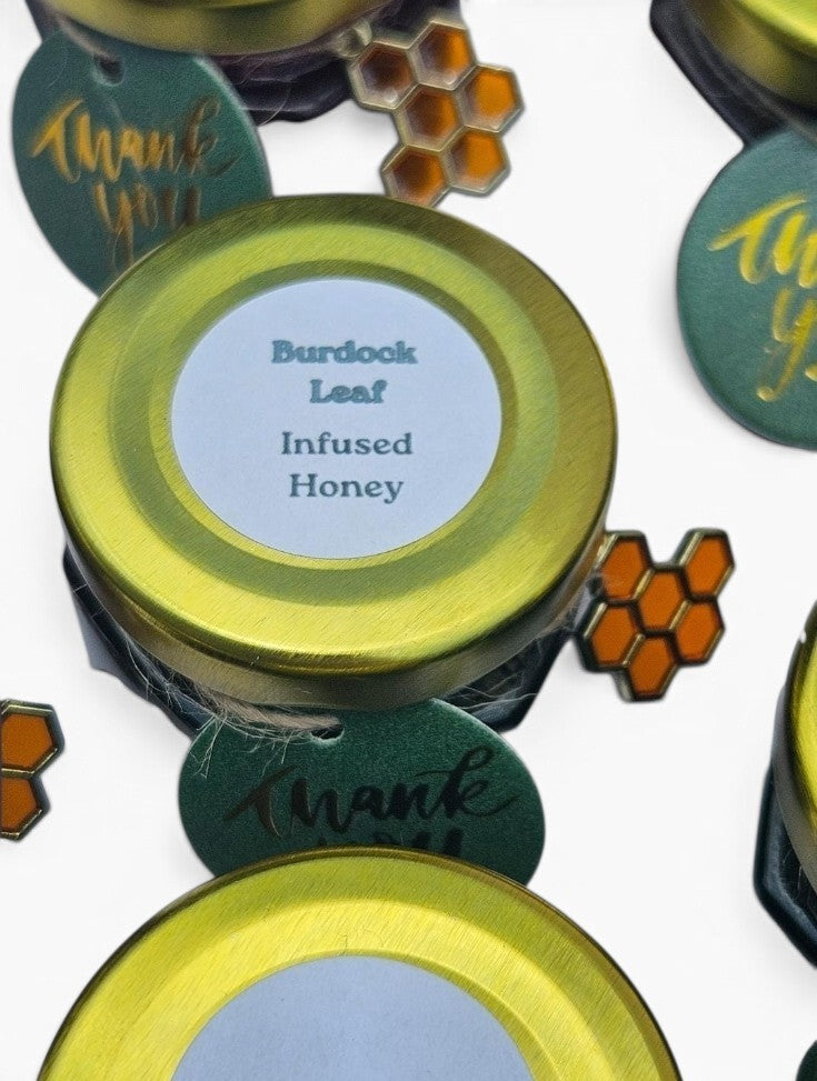A close-up of a jar of infused honey with a gold metal lid and a white label reading 'Burdock Leaf Infused Honey.' Surrounding the jar are decorative green tags with gold script and orange honeycomb motifs on a white background.