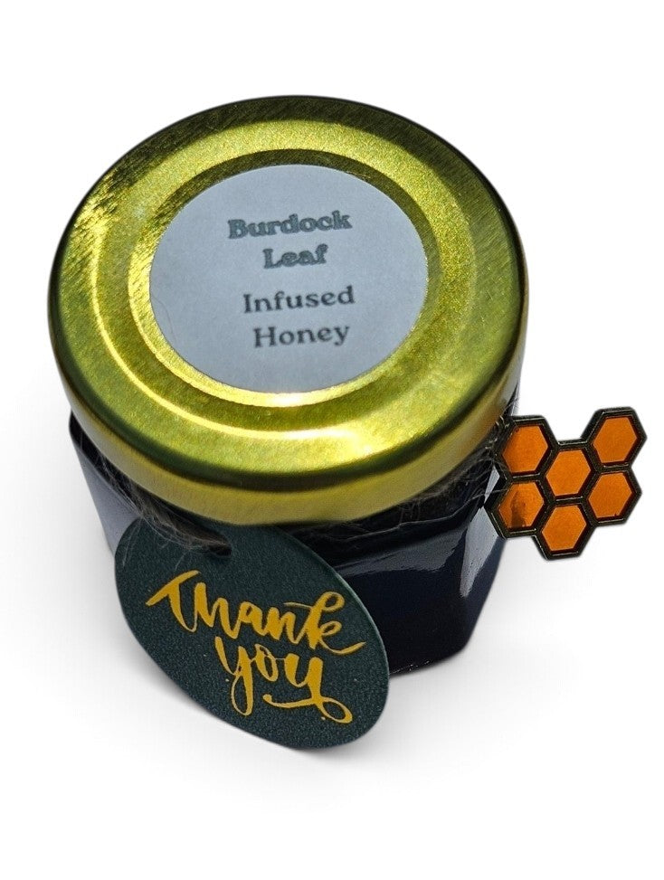A small jar of Burdock Leaf Infused Honey with a shiny gold lid, a black glass body, a dark green 'thank you' hang tag with gold foil script, and an orange honeycomb enamel pin attached to the side, all on a white background.