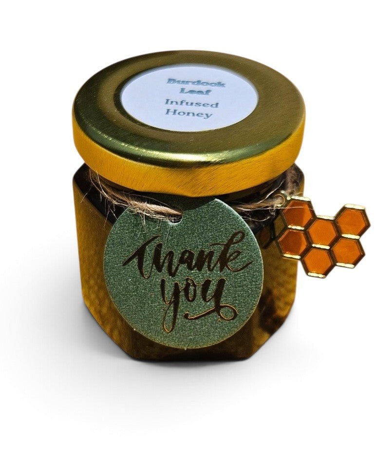 A small jar of burdock leaf infused honey with a gold lid and a round green 'thank you' tag tied to the side, featuring a small honeycomb charm, on a white background.