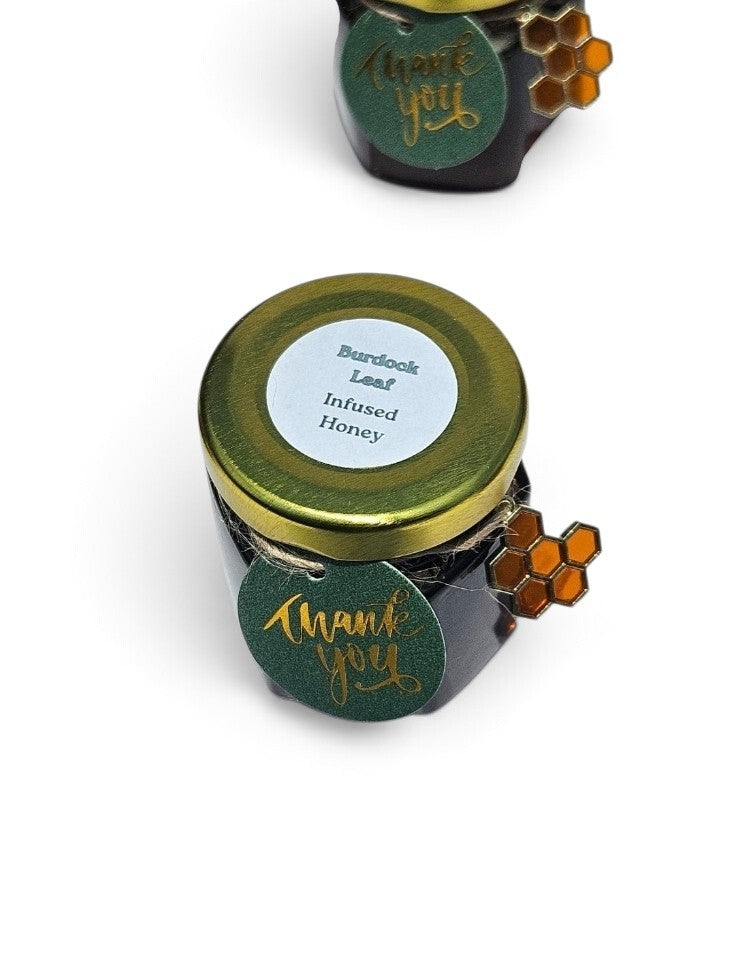 A jar of Burdock Leaf Infused Honey with a gold lid and a green 'Thank You' tag, shown on a white background with a second jar partially visible in the background.