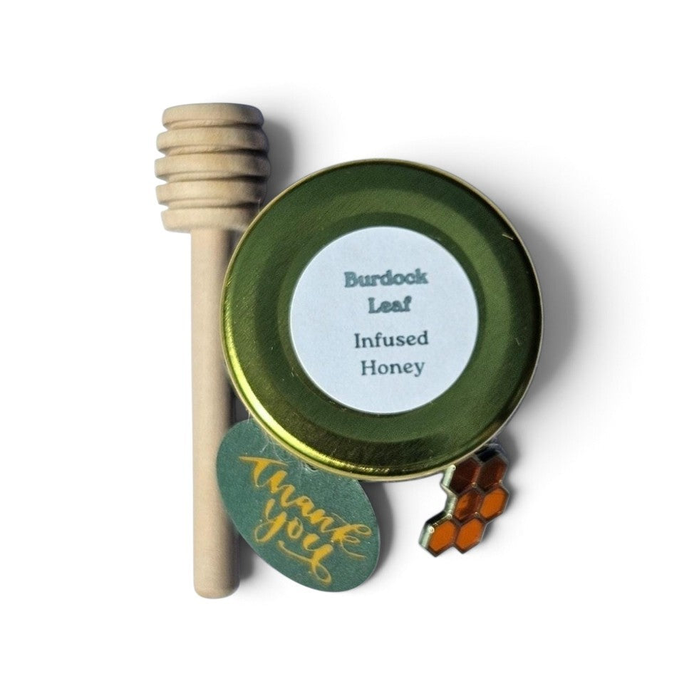 A jar of Burdock Leaf Infused Honey with a wooden honey dipper, a green 'thank you' tag, and a honeycomb charm on a white background.
