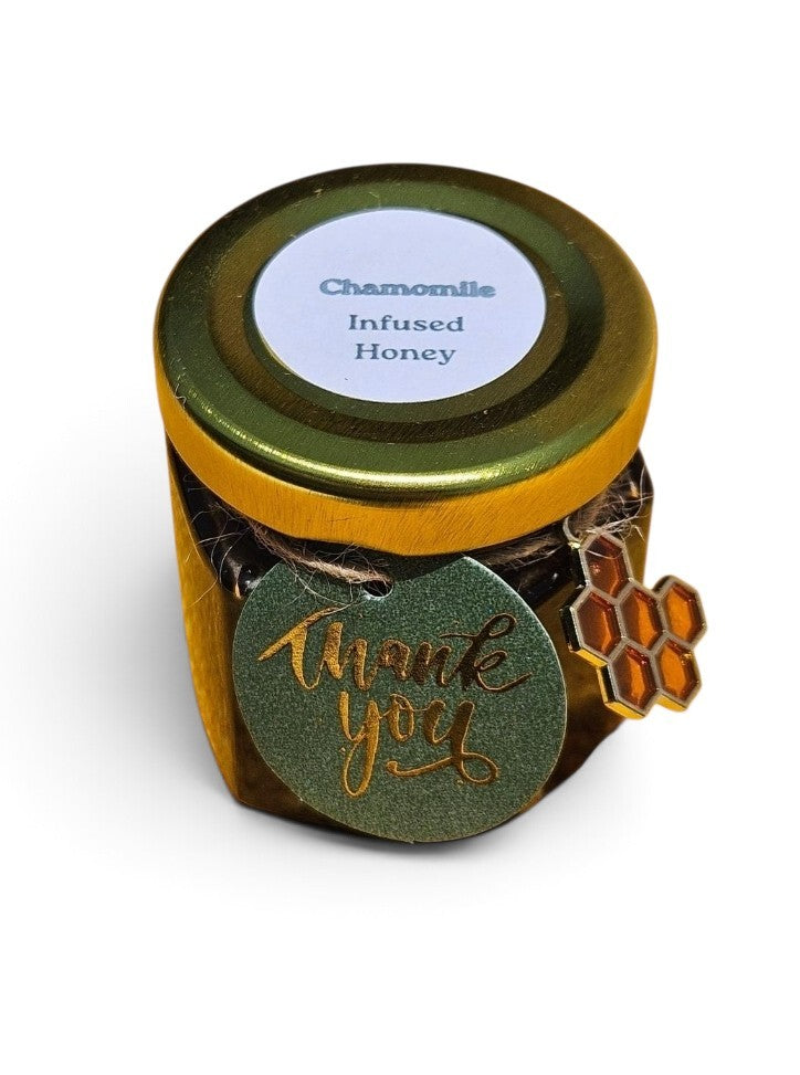 A small jar of chamomile infused honey with a gold lid, a round green 'thank you' tag, and a honeycomb charm, shown on a white background.