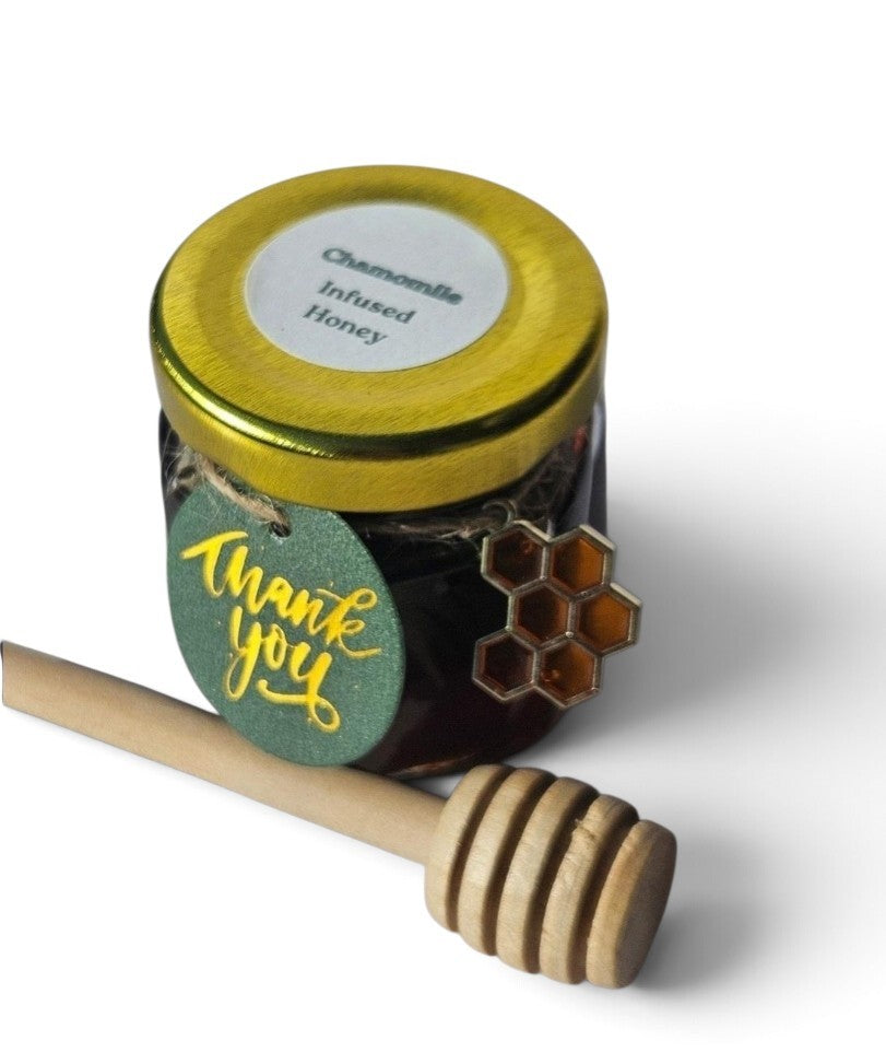 A small glass jar of infused honey with a gold lid and a green 'Thank you' tag, shown with a wooden honey dipper on a white background.