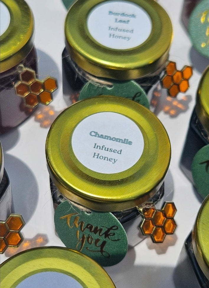 Several small glass jars of infused honey with shiny gold lids are arranged on a white surface. The center jar label reads 'Chamomile Infused Honey,' and nearby tags say 'Thank you.
