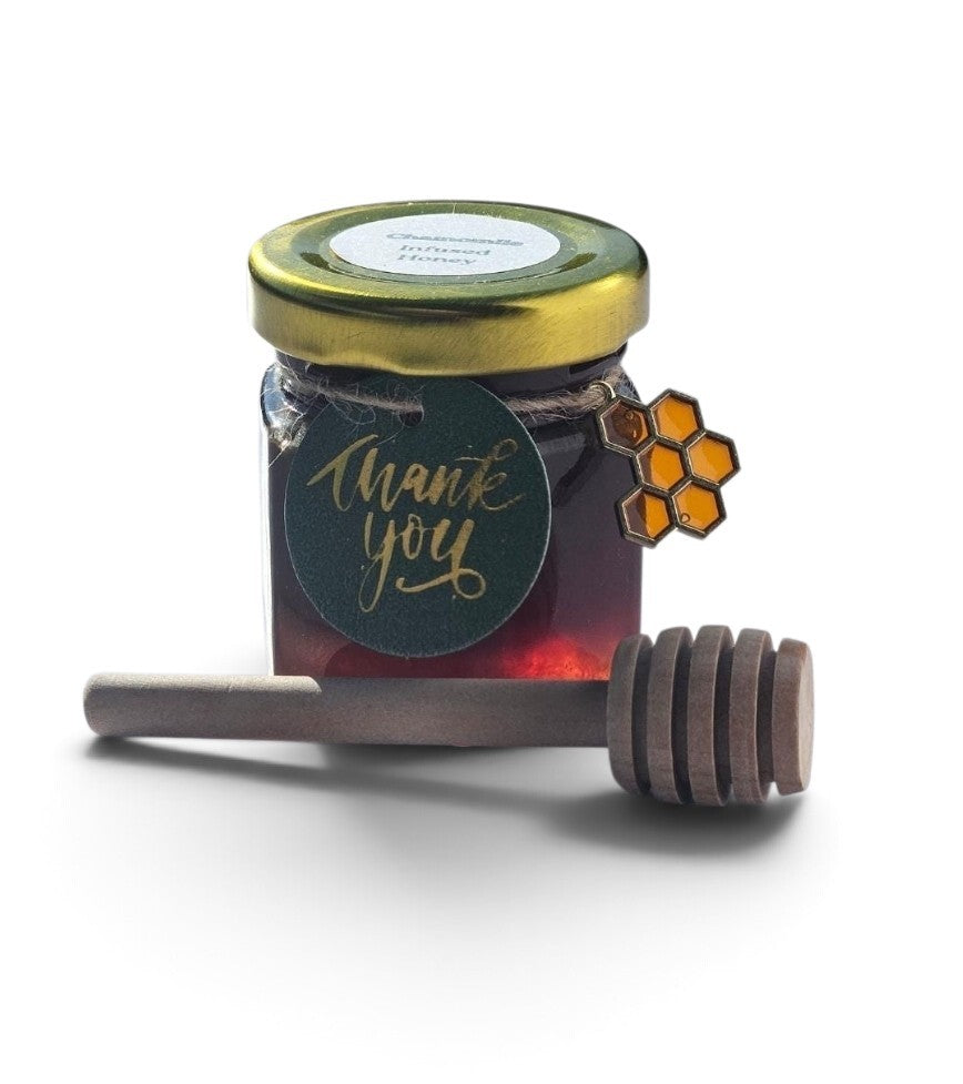 A small glass jar of honey with a gold lid and a dark green round tag reading 'thank you' in gold script, shown with a wooden honey dipper in front on a white background.