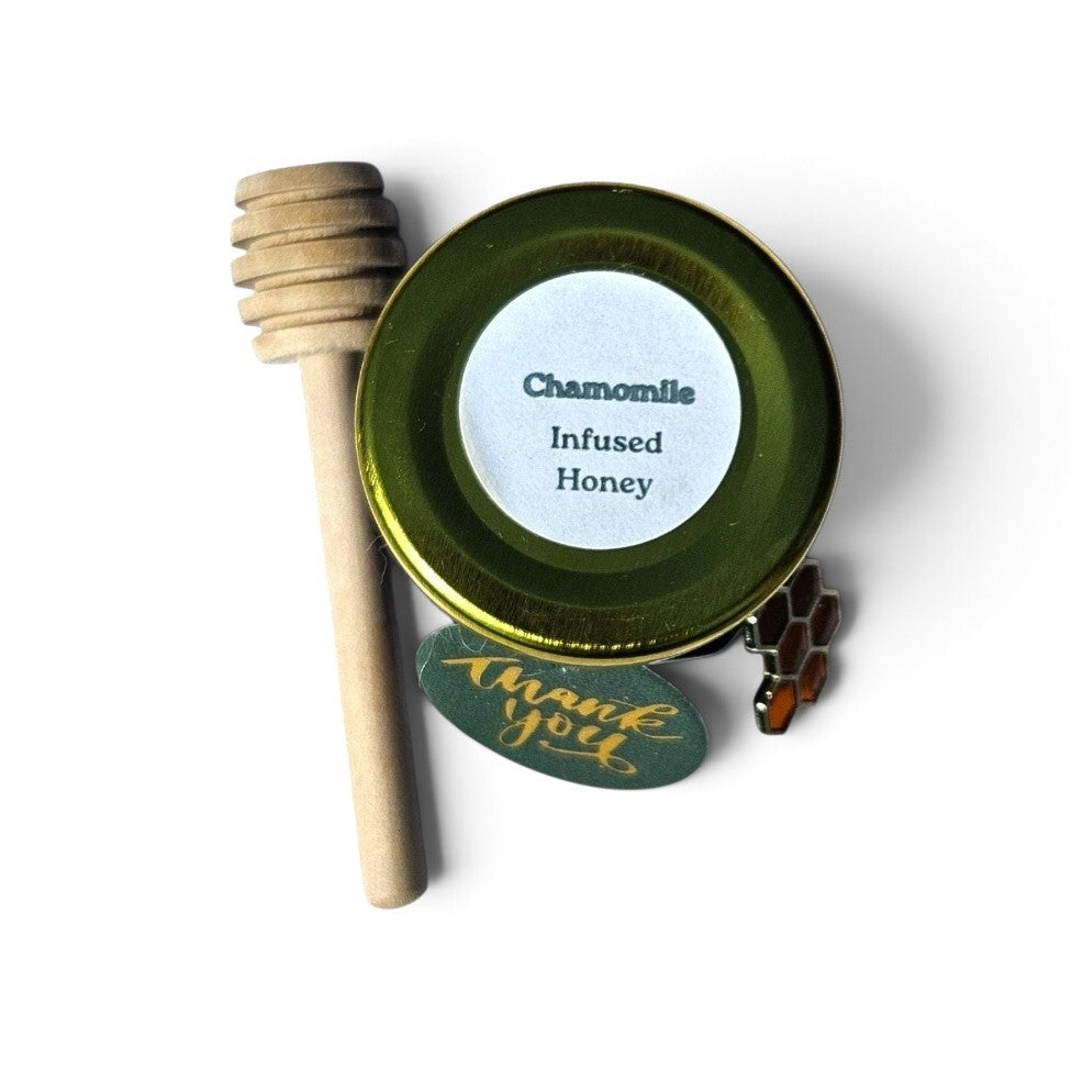 A small round tin of chamomile infused honey with a wooden honey dipper, a small honeycomb charm, and a green 'thank you' tag on a white background.