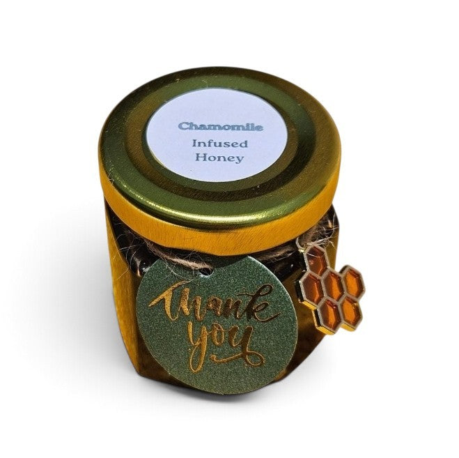 A small glass jar of Chamomile Infused Honey with a gold metal lid, a white circular label, and a green 'thank you' tag with gold foil script. A small honeycomb-shaped charm is attached to the side.