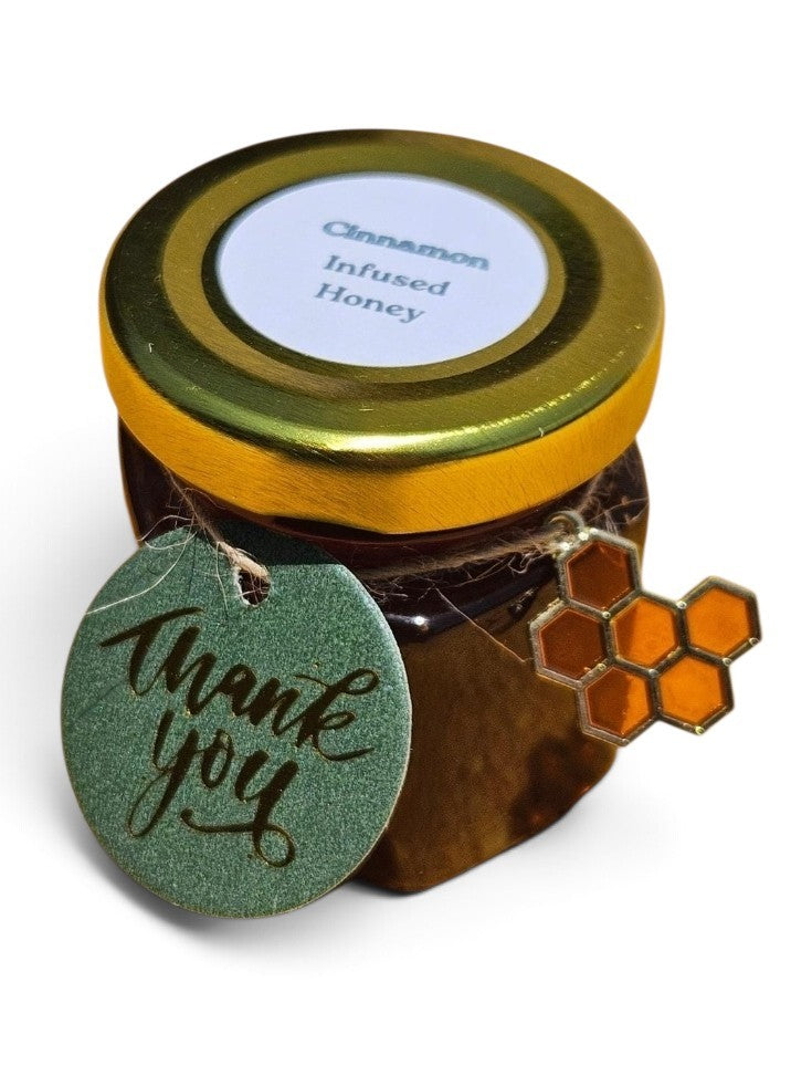 A small jar of cinnamon infused honey with a gold lid and a white label, decorated with a green glittery 'thank you' tag and an orange honeycomb charm, on a white background.