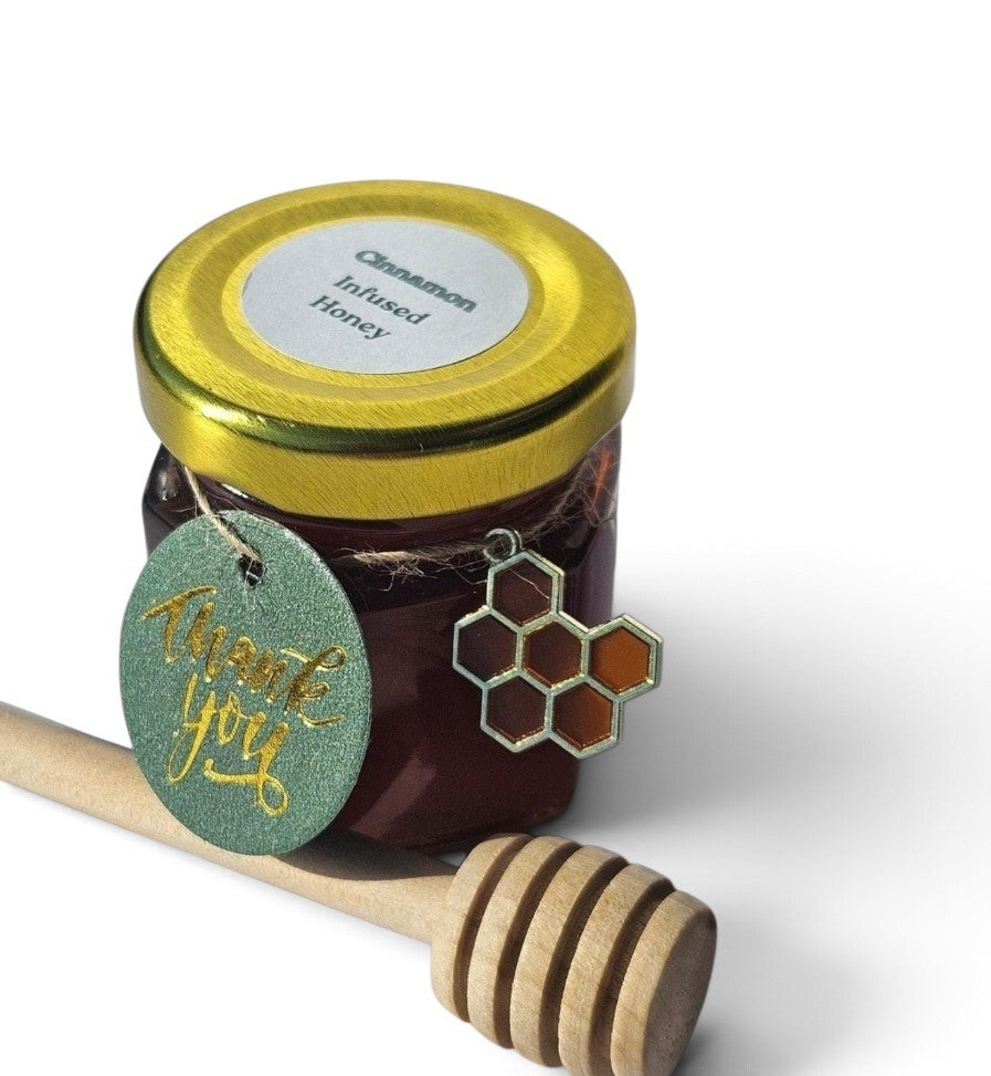 A small glass jar of cinnamon infused honey with a shiny gold lid, a round green 'Thank You' tag, and a honeycomb-shaped charm, shown with a wooden honey dipper on a white background.