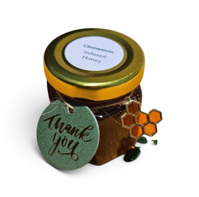 A small glass jar of cinnamon infused honey with a gold lid and a round green 'thank you' tag attached, alongside an orange honeycomb enamel pin, on a white background.