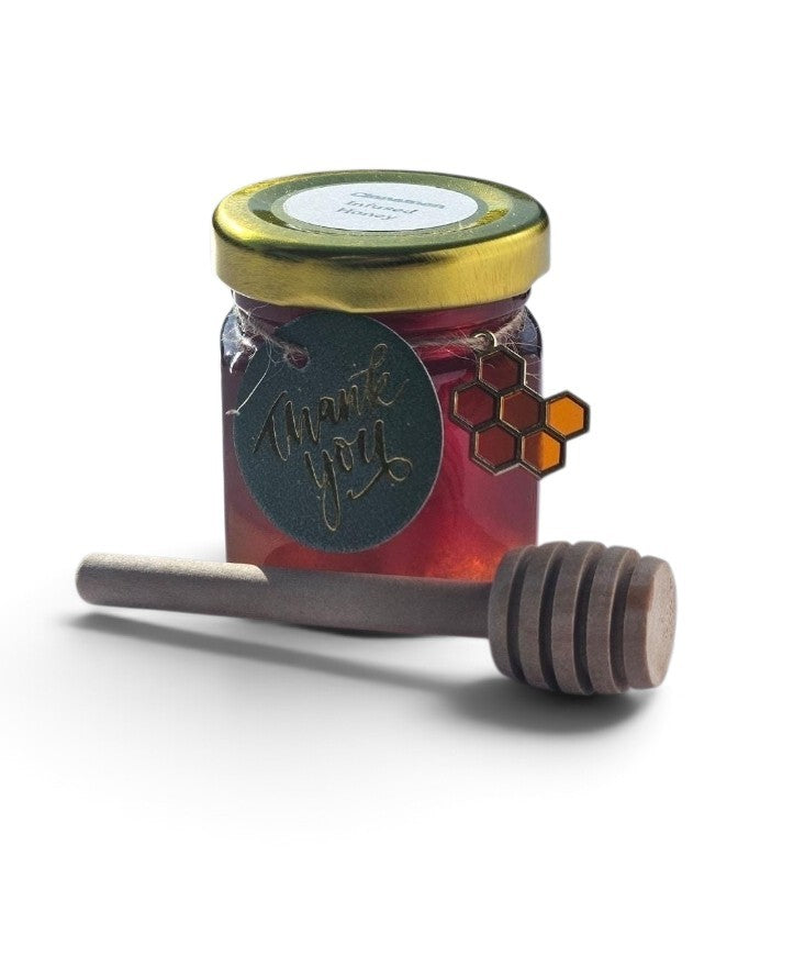 A small glass jar filled with red honey, topped with a gold lid and a round black gift tag that reads 'thank you' in script. A wooden honey dipper rests in front on a white background.