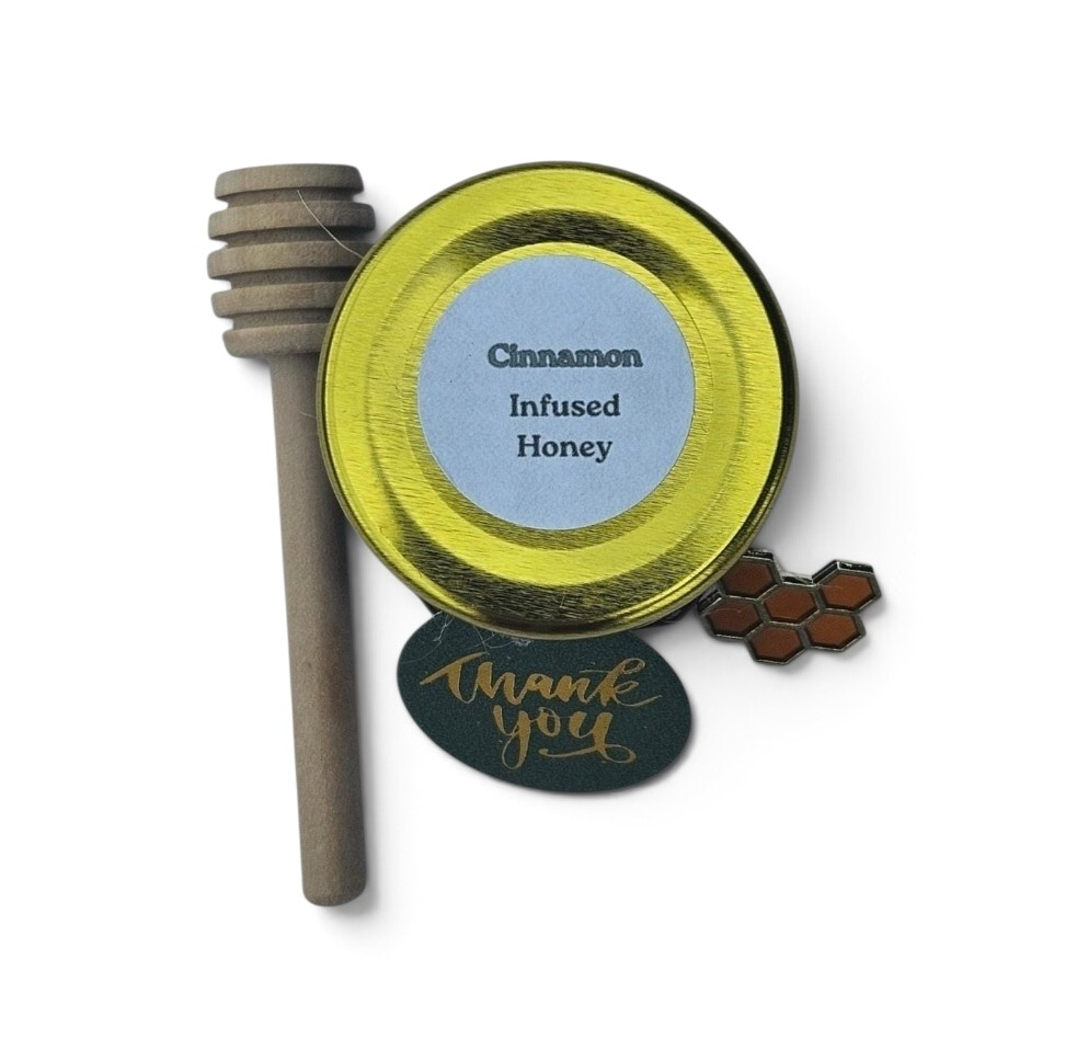 An infused honey sample featuring a round tin of cinnamon infused honey with a wooden honey dipper, a small honeycomb enamel pin, and a dark green 'Thank you' card on a white background.