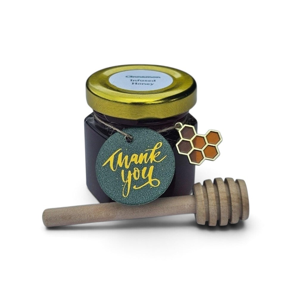 A small jar of cinnamon infused honey with a gold lid, a round teal 'thank you' tag, and a wooden honey dipper on a white background.