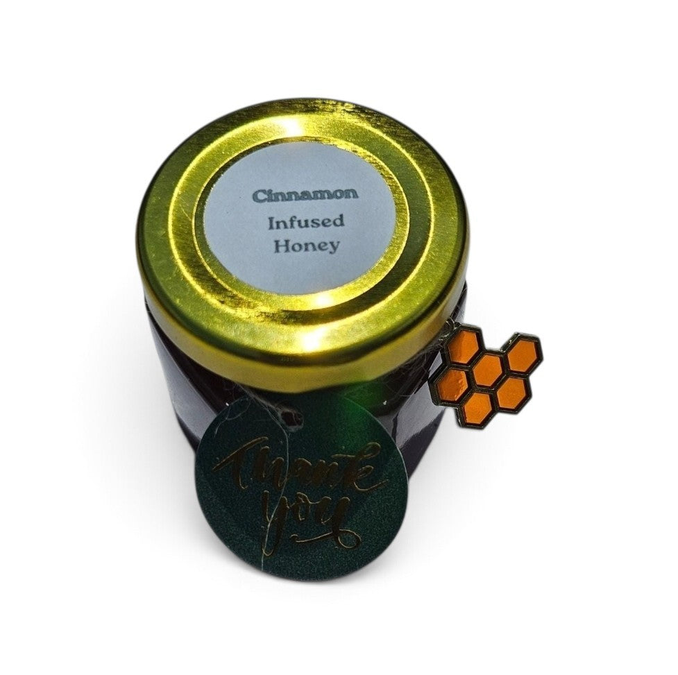 A small jar of cinnamon infused honey with a shiny gold lid and a silver label, shown on a white background with a green round tag and an orange honeycomb charm attached.