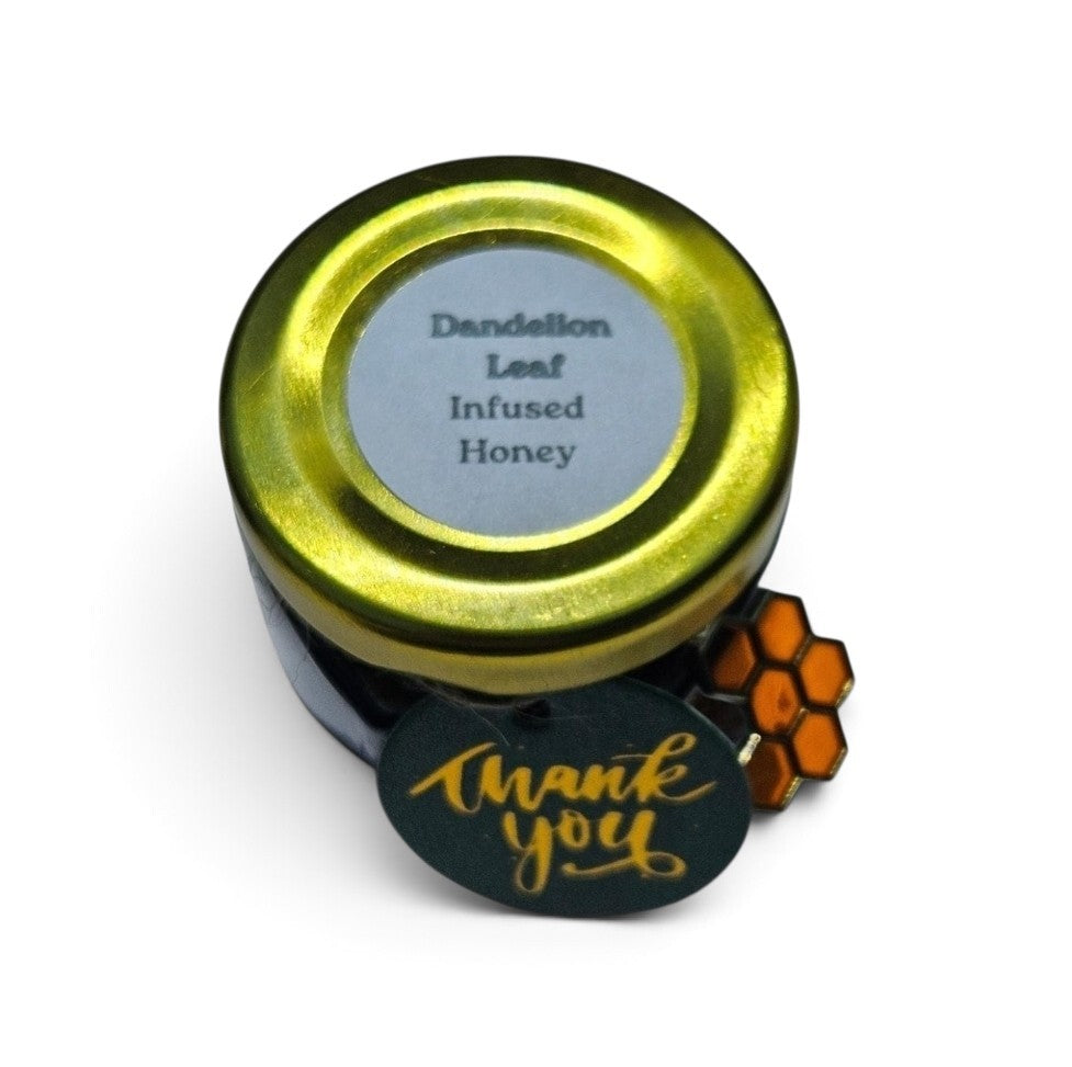 A small jar of Dandelion Leaf Infused Honey with a shiny gold lid and a gray label. A green round tag with gold 'Thank you' script and an orange honeycomb graphic is attached to the jar, set against a plain white background.