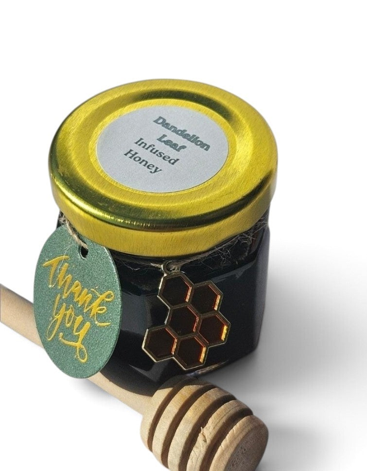 A jar of Dandelion Leaf Infused Honey with a gold lid, a green 'Thank You' tag, and a wooden honey dipper on a white background.