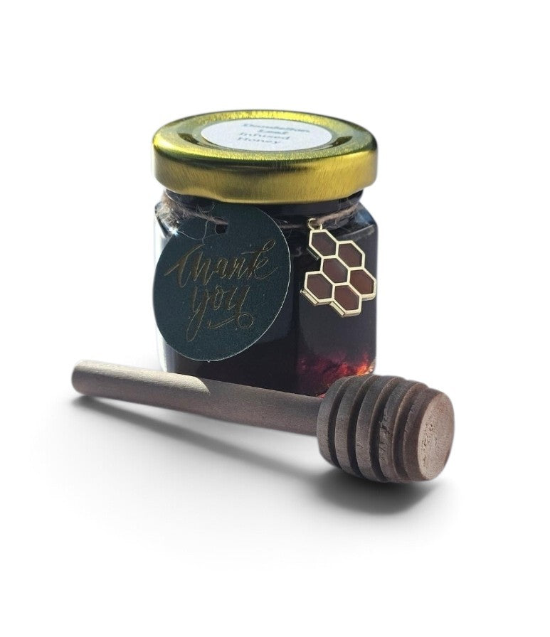 A small glass jar of dark honey with a gold metal lid and a round black 'thank you' tag, shown with a wooden honey dipper in front on a white background.