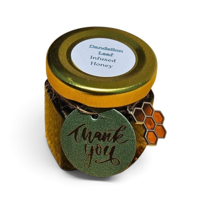 A small jar of Dandelion Leaf Infused Honey with a gold lid, a white label, and a green glittery 'thank you' tag with a honeycomb charm, shown on a white background.