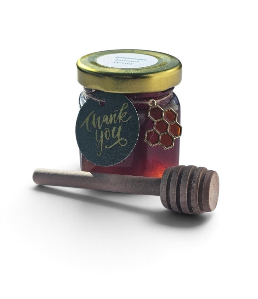 A small glass jar of echinacea infused honey with a gold metal lid, a dark green round 'thank you' tag, and a wooden honey dipper resting in front, photographed on a clean white background.