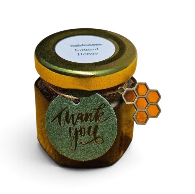 A small jar of Echinacea Infused Honey with a gold lid, featuring a green glittery 'thank you' tag and a honeycomb-shaped tag on the side, set against a plain white background.