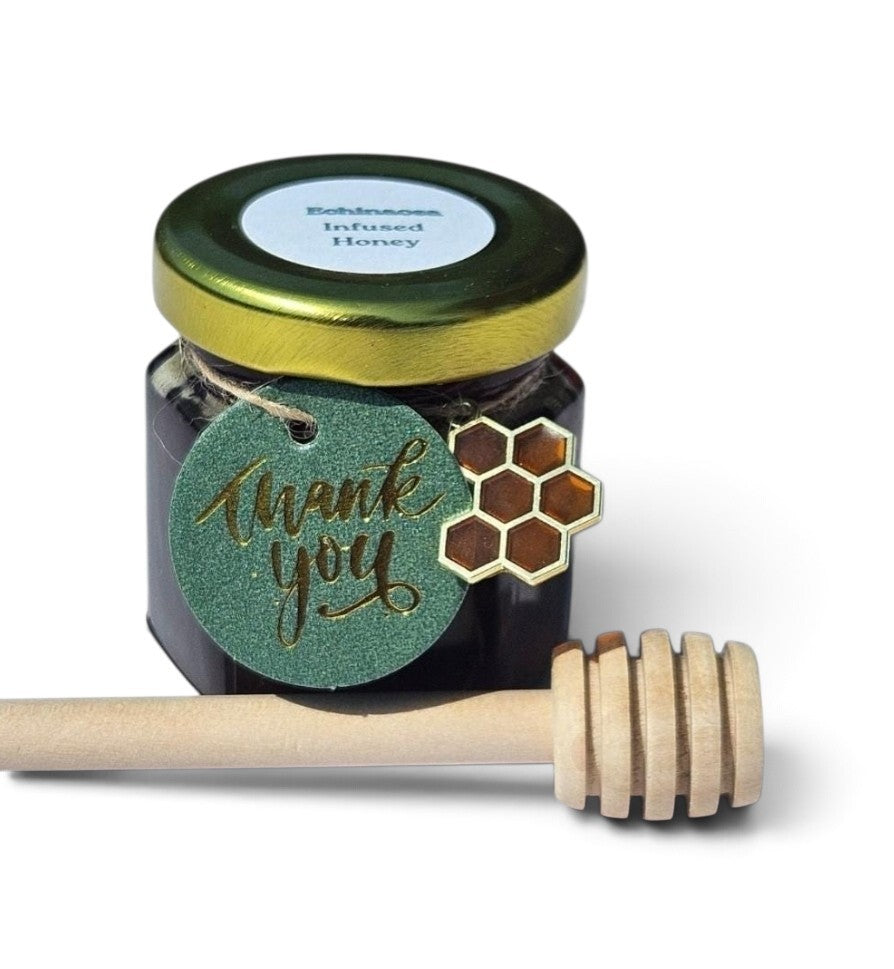A jar of echinacea infused honey with a gold lid and a green 'thank you' tag, shown with a wooden honey dipper on a white background.