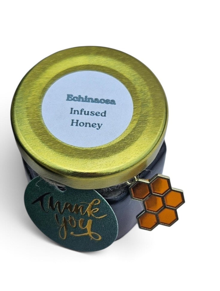A jar of Echinacea Infused Honey with a shiny gold lid, a white circular label, and a green 'thank you' tag with gold foil script and an orange honeycomb charm, on a white background.