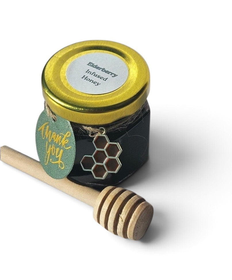 A small jar of elderberry infused honey with a gold lid and a green 'thank you' tag, shown with a wooden honey dipper on a white background.