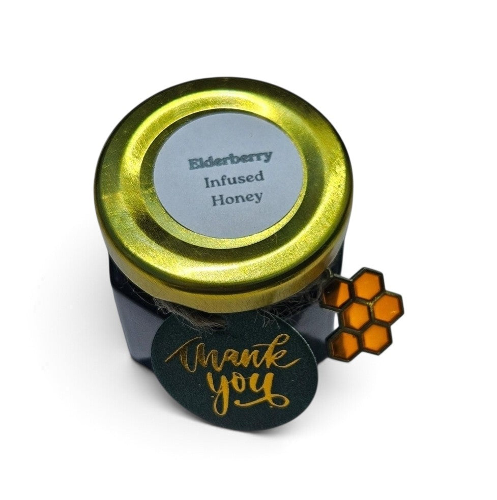 A small jar of elderberry infused honey with a gold metal lid, a white label reading 'Elderberry Infused Honey,' and a dark green round tag that says 'Thank you' in gold script. A small orange honeycomb charm is attached on the side.