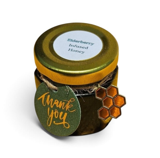 A small jar of elderberry infused honey with a gold metal lid, a white circular label, a green 'thank you' tag, and a honeycomb-shaped charm on a white background.
