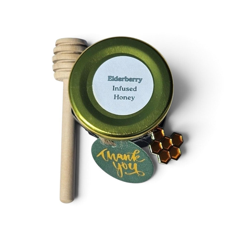 A small round tin of elderberry infused honey with a wooden honey dipper, a green 'Thank You' tag, and a honeycomb charm on a white background.