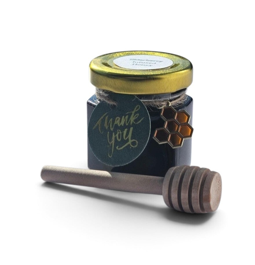 A sample sized jar of elderberry honey with a gold lid and a round dark green 'thank you' tag, shown with a wooden honey dipper on a white background.
