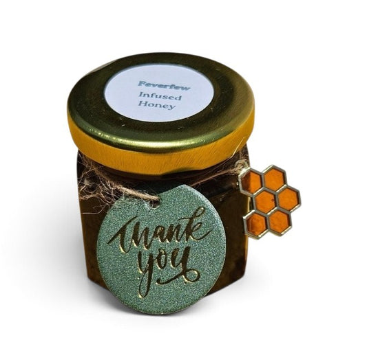 A small jar of Feverfew Infused Honey with a gold lid, a round green 'thank you' tag, and an orange honeycomb charm, shown on a white background.