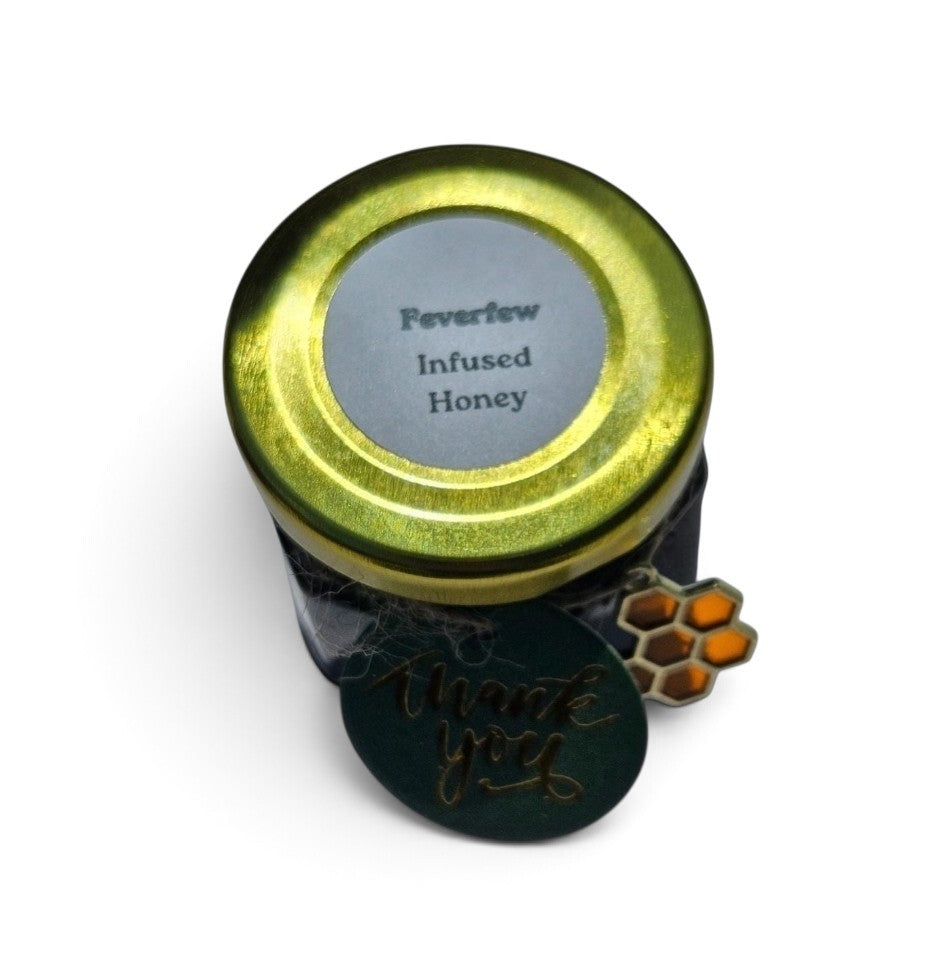 A jar of Feverfew Infused Honey with a metallic gold lid, a dark glass body, and a round dark green tag that reads 'thank you' in gold script, shown on a white background.