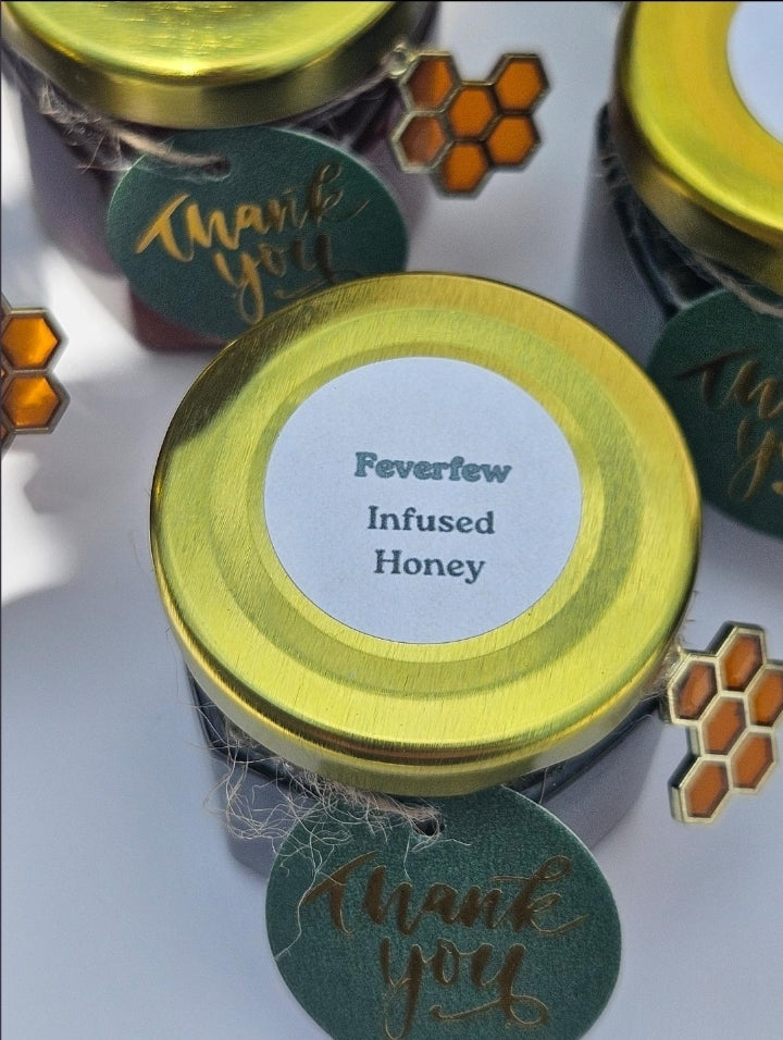A close-up of small glass jars filled with honey, each topped with a shiny gold lid and a round white label reading 'Feverfew Infused Honey.' Dark green hanging tags with gold script say 'thank you,' and decorative honeycomb graphics appear around the jars on a light background.