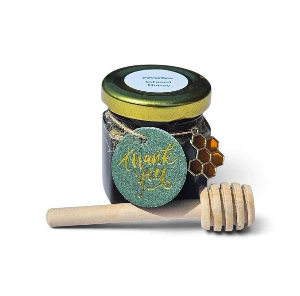 A small glass jar of feverfew honey with a gold lid, a round teal 'thank you' tag with gold script, a small honeycomb charm, and a wooden honey dipper on a white background.