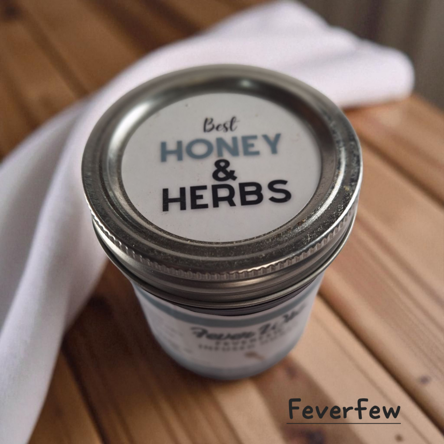 Top view of 8 oz half-pint jar of raw Feverfew Infused Honey on a rustic wooden surface, herbal sweetener for tea, baking, and traditional herbal uses.