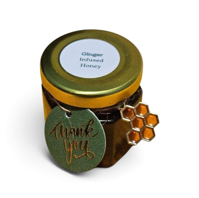 A small glass jar of ginger infused honey with a gold metal lid and a round green 'thank you' tag hanging from the side, featuring a small honeycomb charm, on a white background.