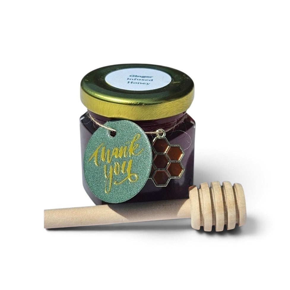 A jar of ginger infused honey with a gold lid and a green 'thank you' tag, shown with a wooden honey dipper on a white background.